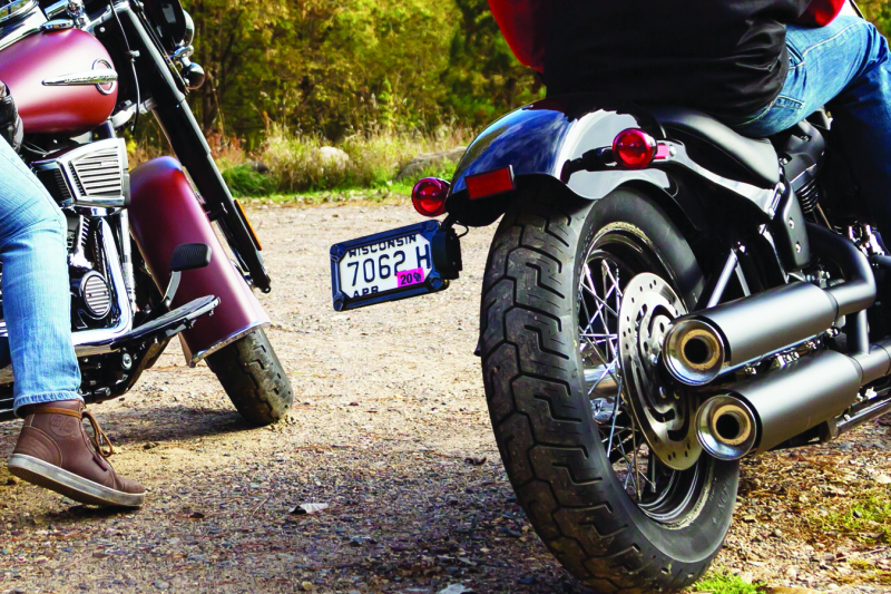 Kuryakyn Nova side mount license plate frame for Harley Davidson in gloss black, with mounting hardware visible.